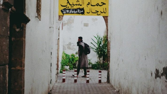 Man walking through a rustic alleyway with Arabic signage, surrounded by white walls and vegetation.